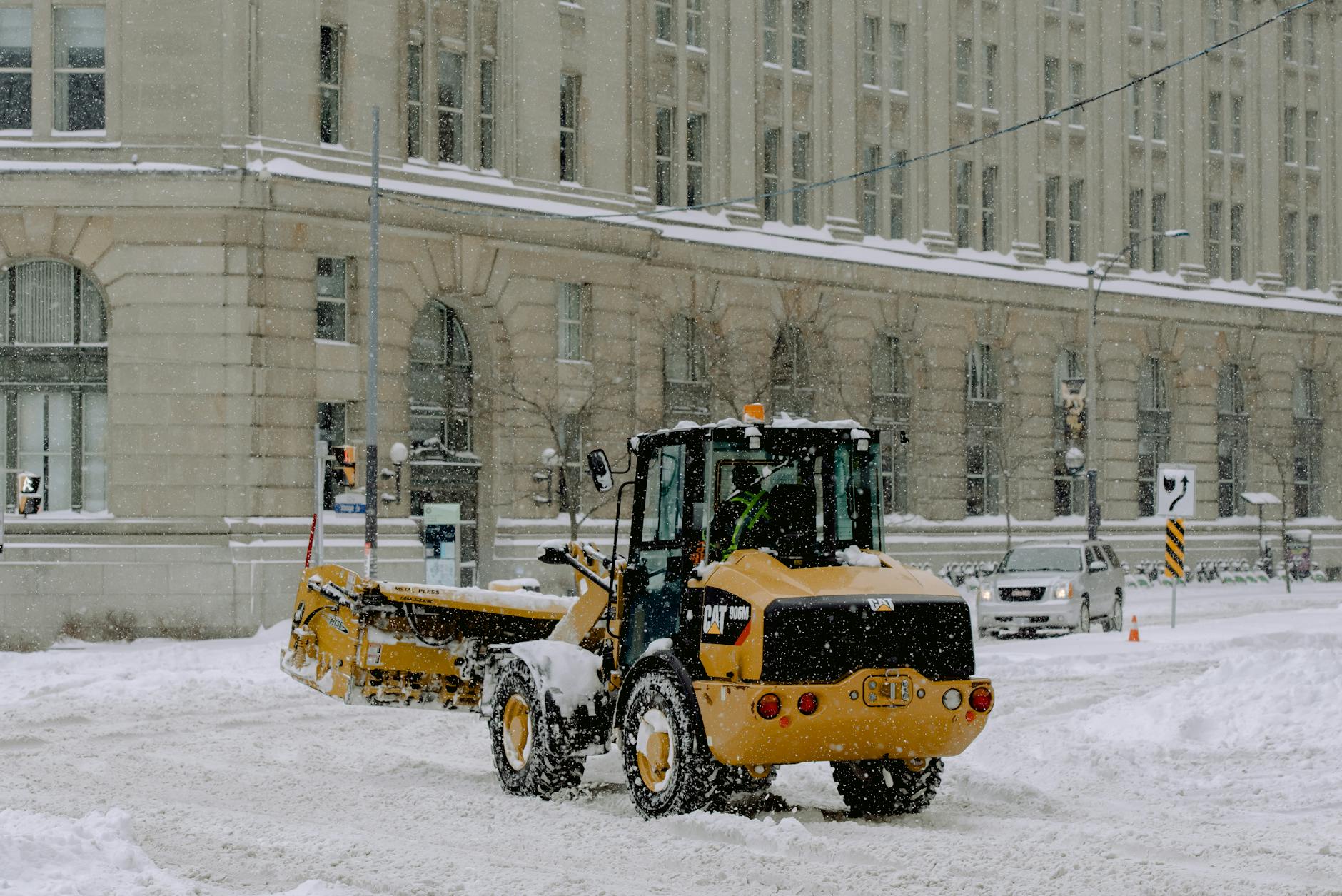 A snowplow clearing snow on a snowy city street with a historical building backdrop.
